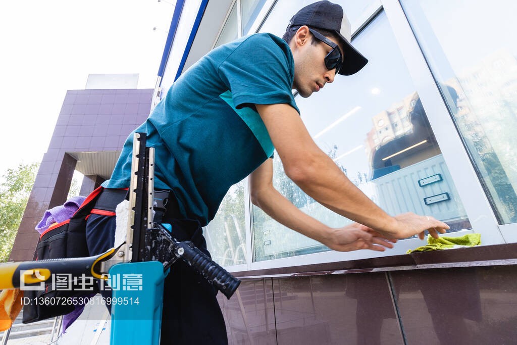 一名穿着工作服的专业清洁服务的年轻男性工人正在清洗大楼正面的窗户玻璃。应要求清洁陈列柜